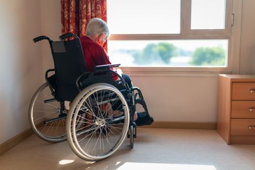An older woman with white hair, alone in her house while looking outside through the window - protect your estate from nursing home costs concept.